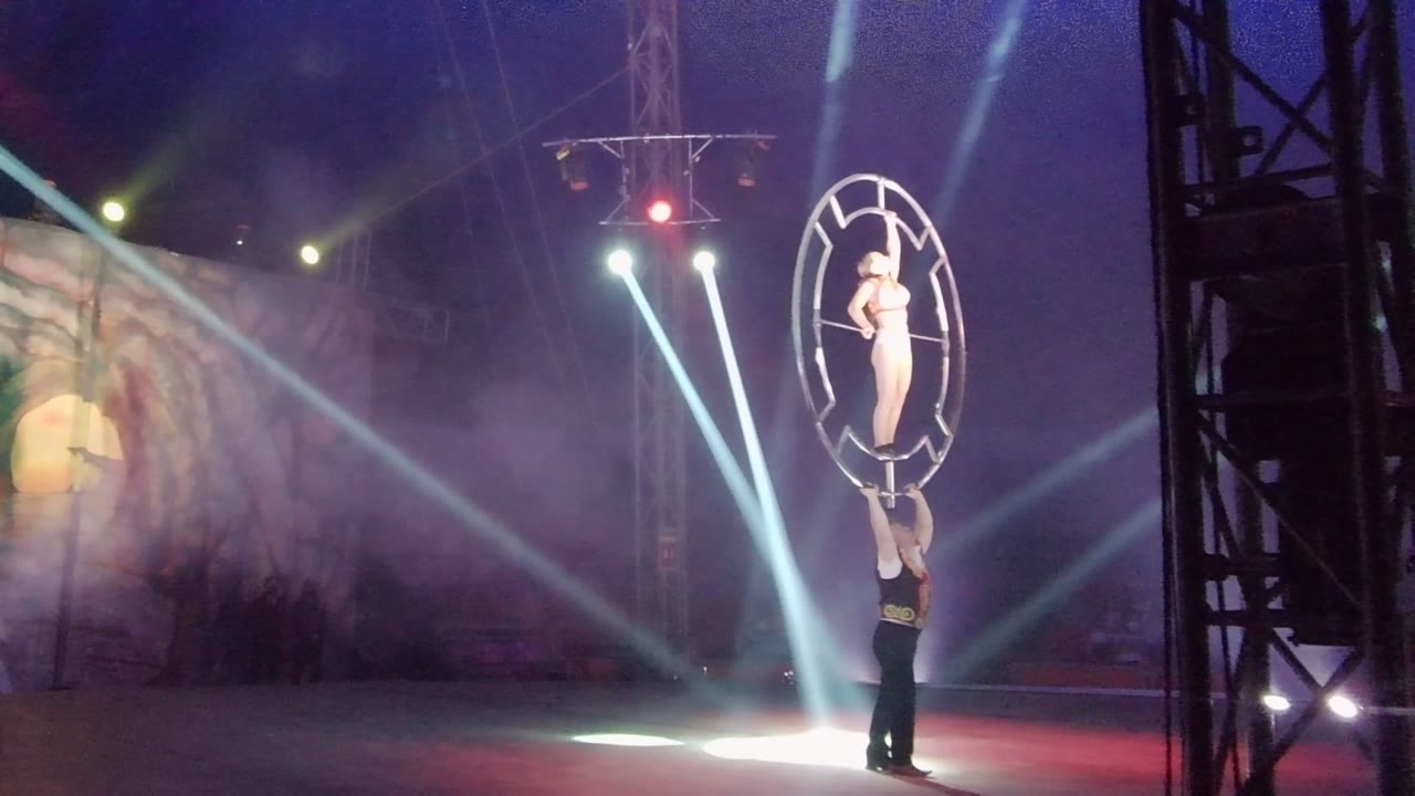 Aerial hoop performer held above the stage with smoke and spotlights behind.