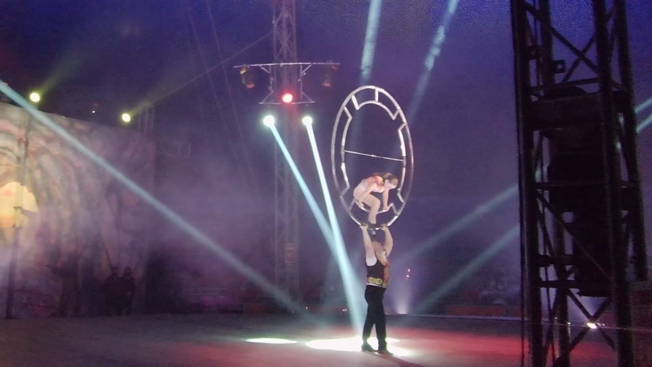 Acrobats balancing near an aerial hoop under crossing stage lights.