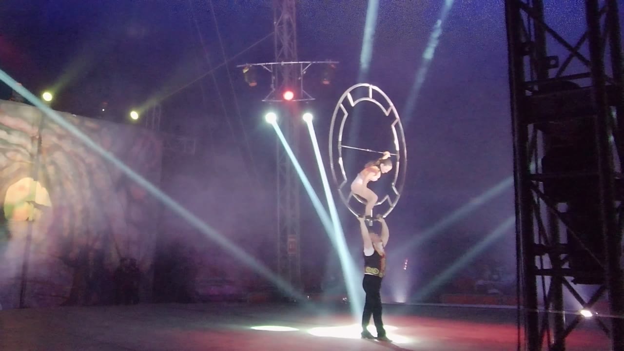 In San Jose del Cabo, Mexico, the female acrobat leans across the aerial hoop as the male performer supports her from below amid bright spotlights and smoky blue-purple stage light.