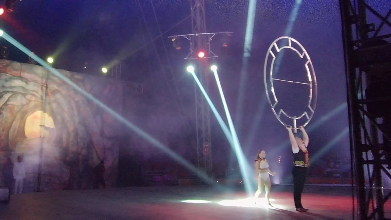 In San Jose del Cabo, Mexico, two performers stand on a smoky stage as one lifts a large circular frame near blue-white light beams and a moon-and-trees backdrop.