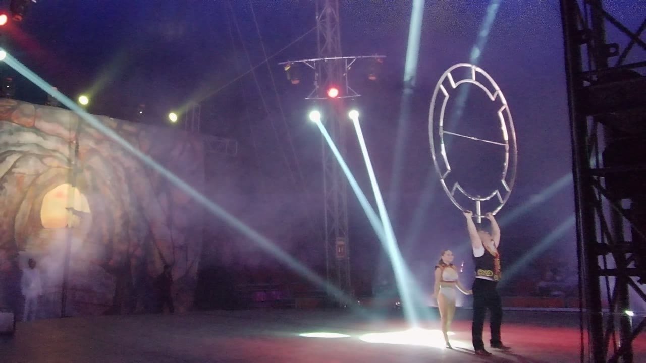 Performer holds a circular hoop frame while another performer stands nearby in stage light.