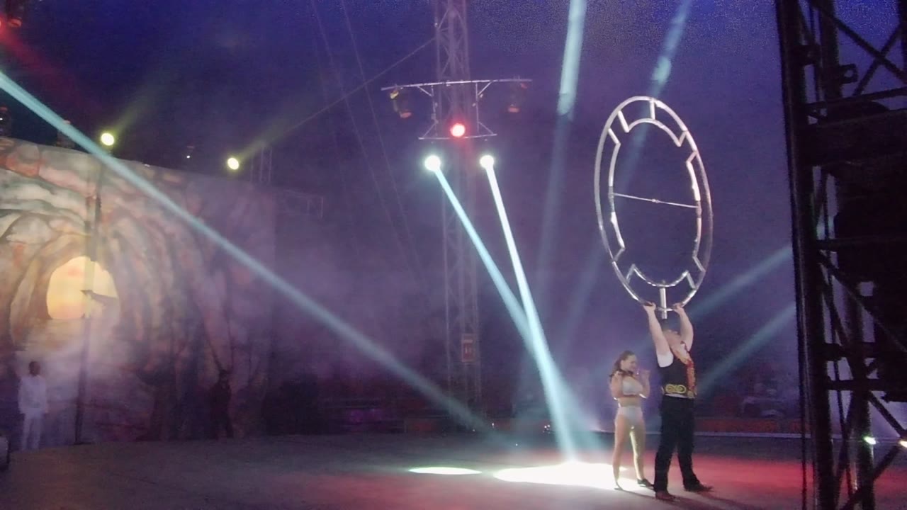 The circular frame remains high on the right side of the San Jose del Cabo stage while another performer waits to the left under cool beams and smoky colored light.