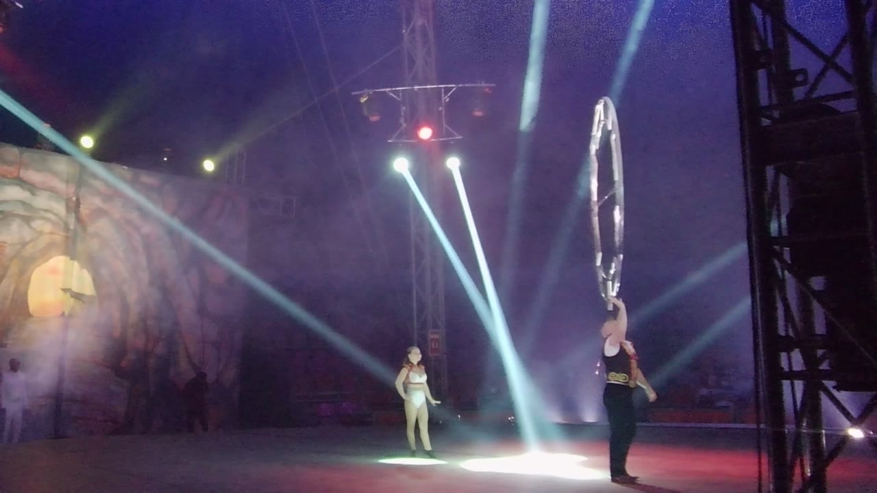 Two performers stand on a smoky stage in San Jose del Cabo, Mexico, with a large circular prop at right, bright floor light near center, and a painted backdrop at left.