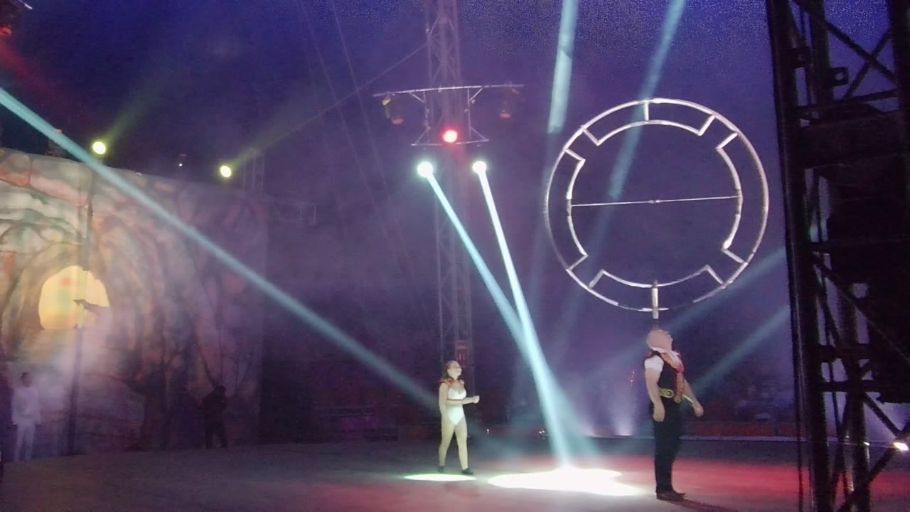 Performer in dark clothing holds a large circular prop while another performer stands in white light