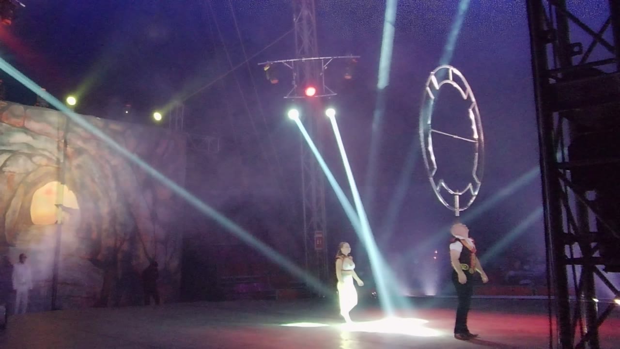 The circular prop is raised higher above the performer on the right as the second performer remains near the center spotlight, with smoke and diagonal light beams across the stage.