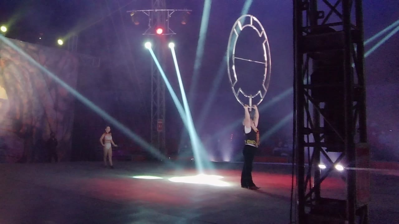 In San Jose Del Cabo BCS Mexico, a stage performer at right holds a large circular frame overhead while a distant dancer stands left of center under blue-white beams and haze.
