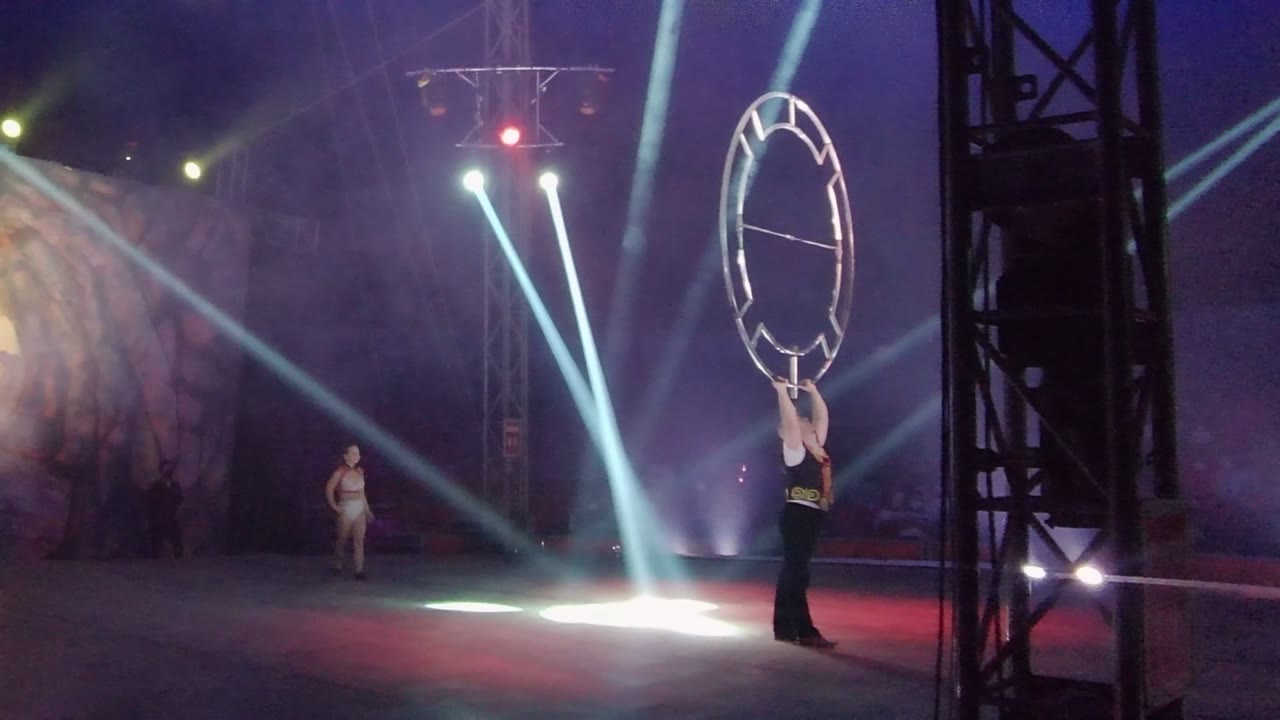 Dancer walks closer while the ring performer remains at right under bright stage beams.
