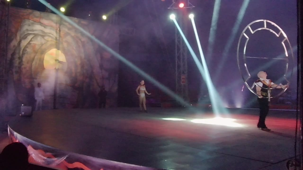 Performer leans back while moving a large circular prop on a smoky stage.