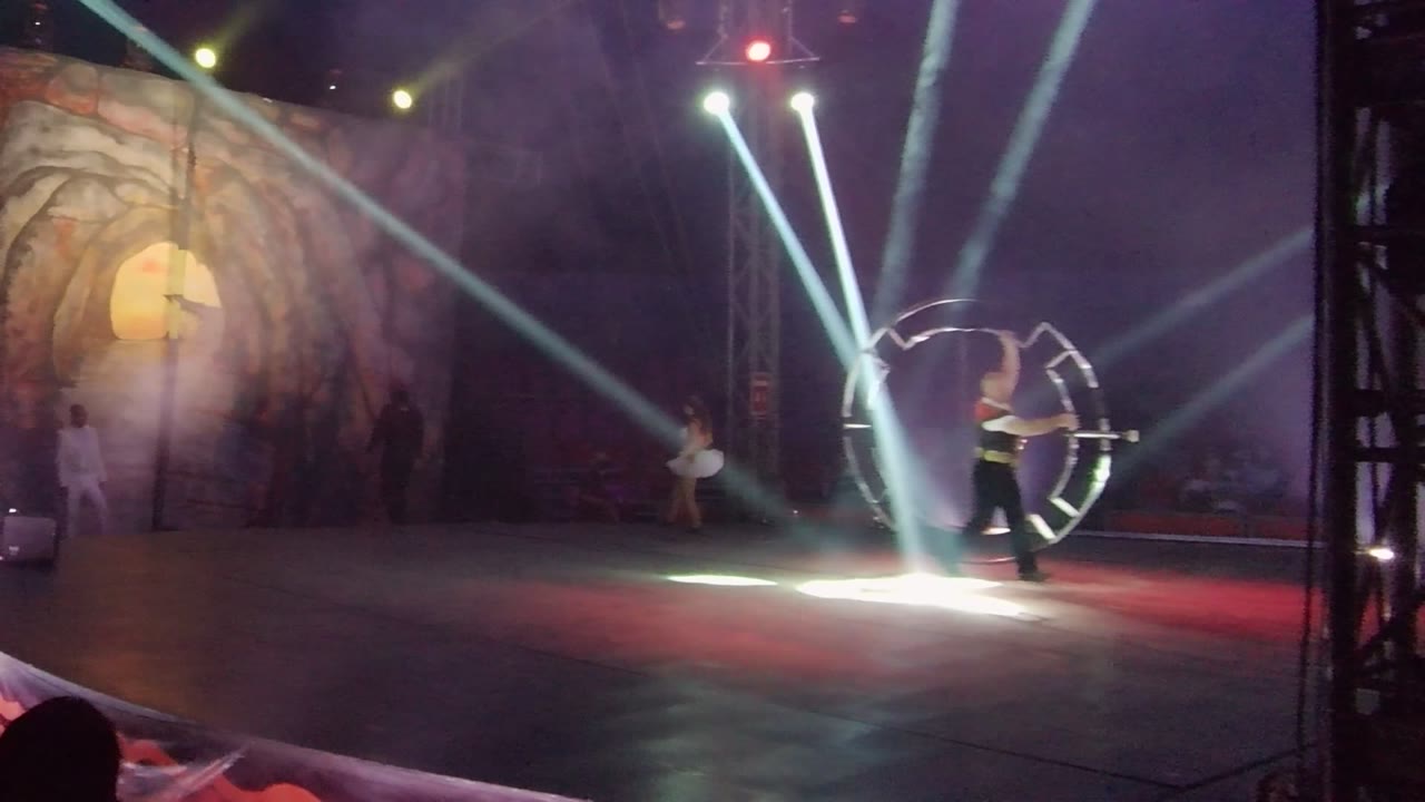 In San Jose del Cabo BCS Mexico, an acrobat stands near a large hoop while white spotlights cut through haze across a cave-like stage backdrop.