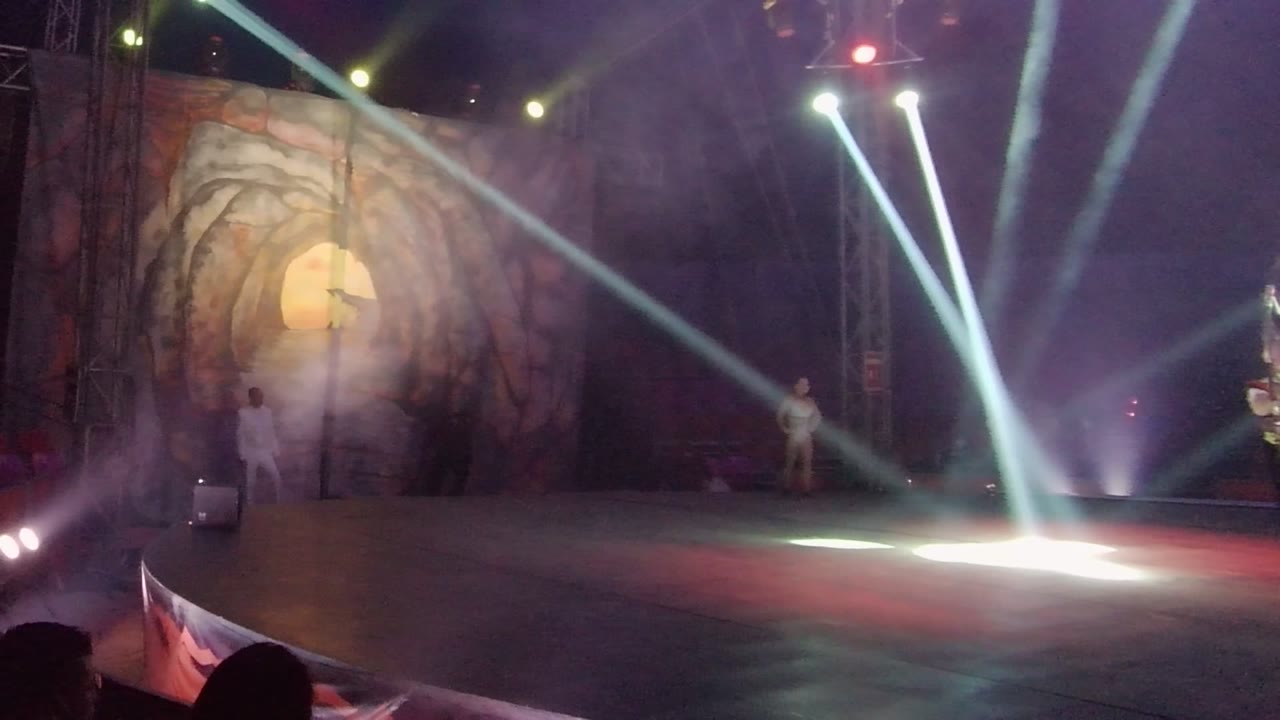 A white-costumed performer stands at the right side of the San Jose del Cabo BCS Mexico stage while the hoop act has moved partly out of frame and spotlights pool on the floor.