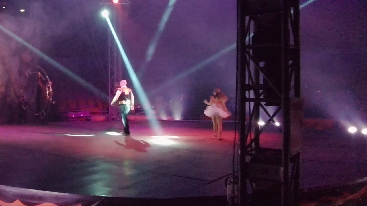 Dancer in dark clothing and dancer in a white dress perform near a large circular hoop.
