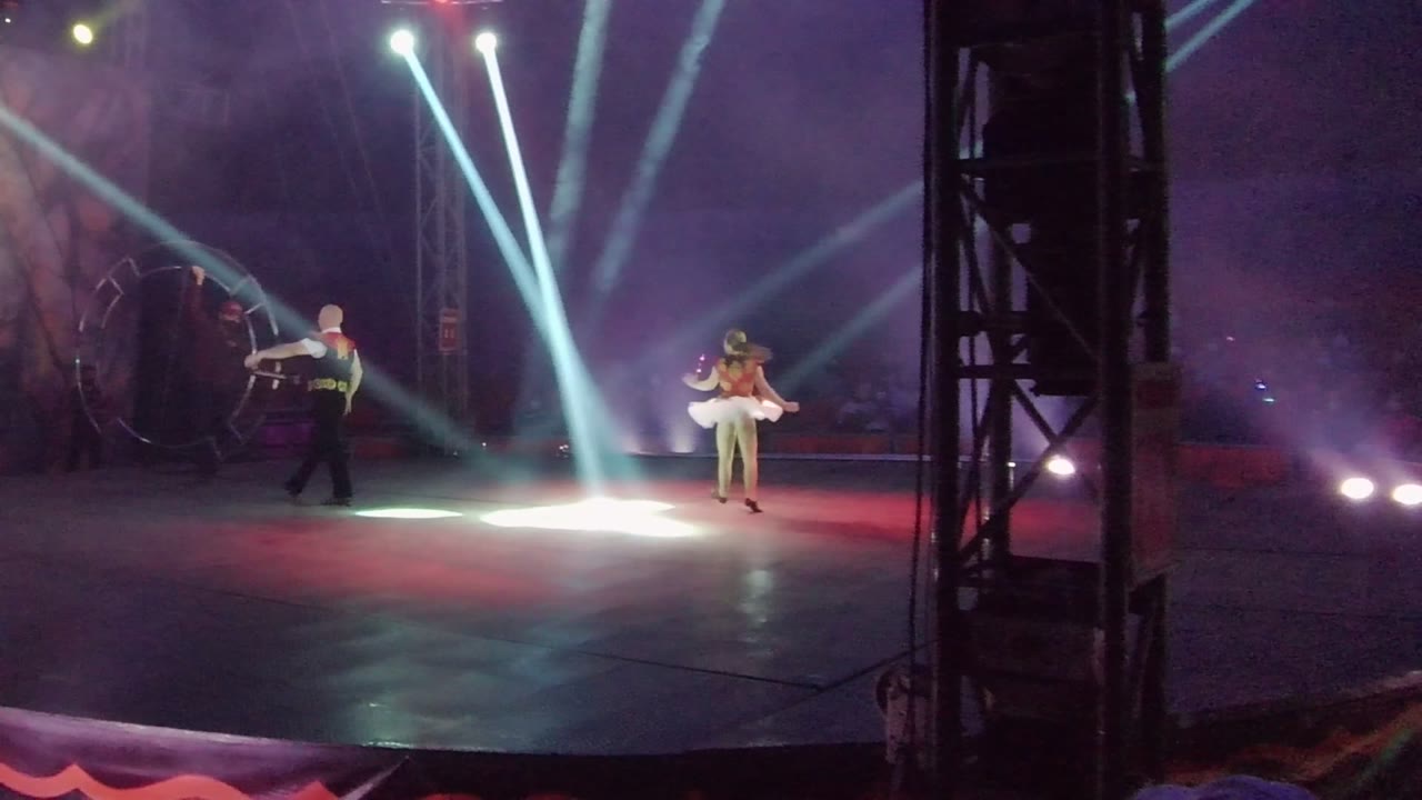 In San Jose del Cabo BCS Mexico, the dark-clad dancer is closer to the hoop at left while the dancer in a white dress is at right beneath crossing white beams and purple smoke.