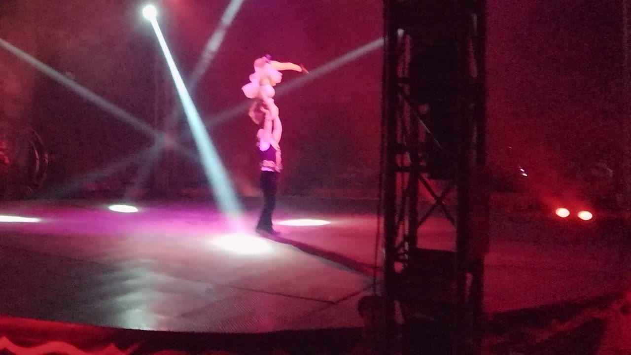 Two acrobats perform an overhead lift on a dark stage in San Jose del Cabo, Mexico, with white beams crossing a red-purple haze and a black truss near the right side.