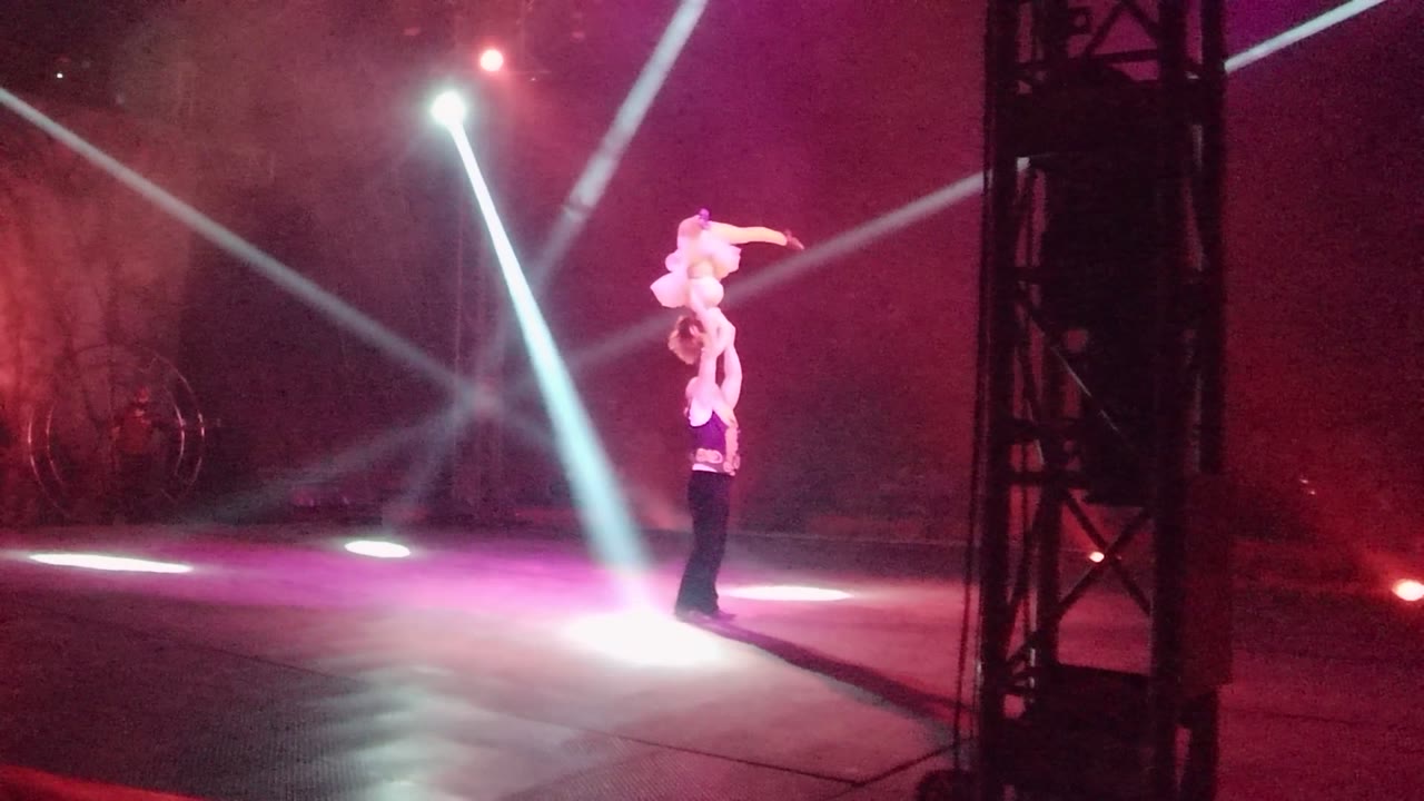 Acrobatic duo stays in a lifted pose beneath bright white stage lights.
