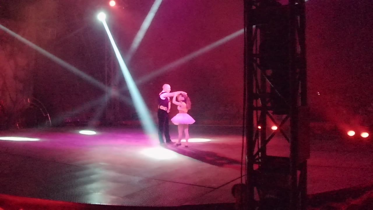 In San Jose del Cabo, Mexico, a male dancer and female dancer stand together on a stage as white spotlights cross through red haze and a black truss borders the right side.