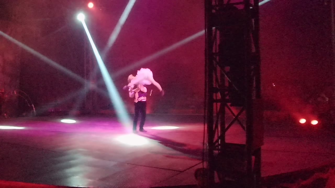 In San Jose del Cabo, Mexico, the male dancer stands alone in the spotlight area while holding the female dancer high across his shoulders against a red-lit stage background.