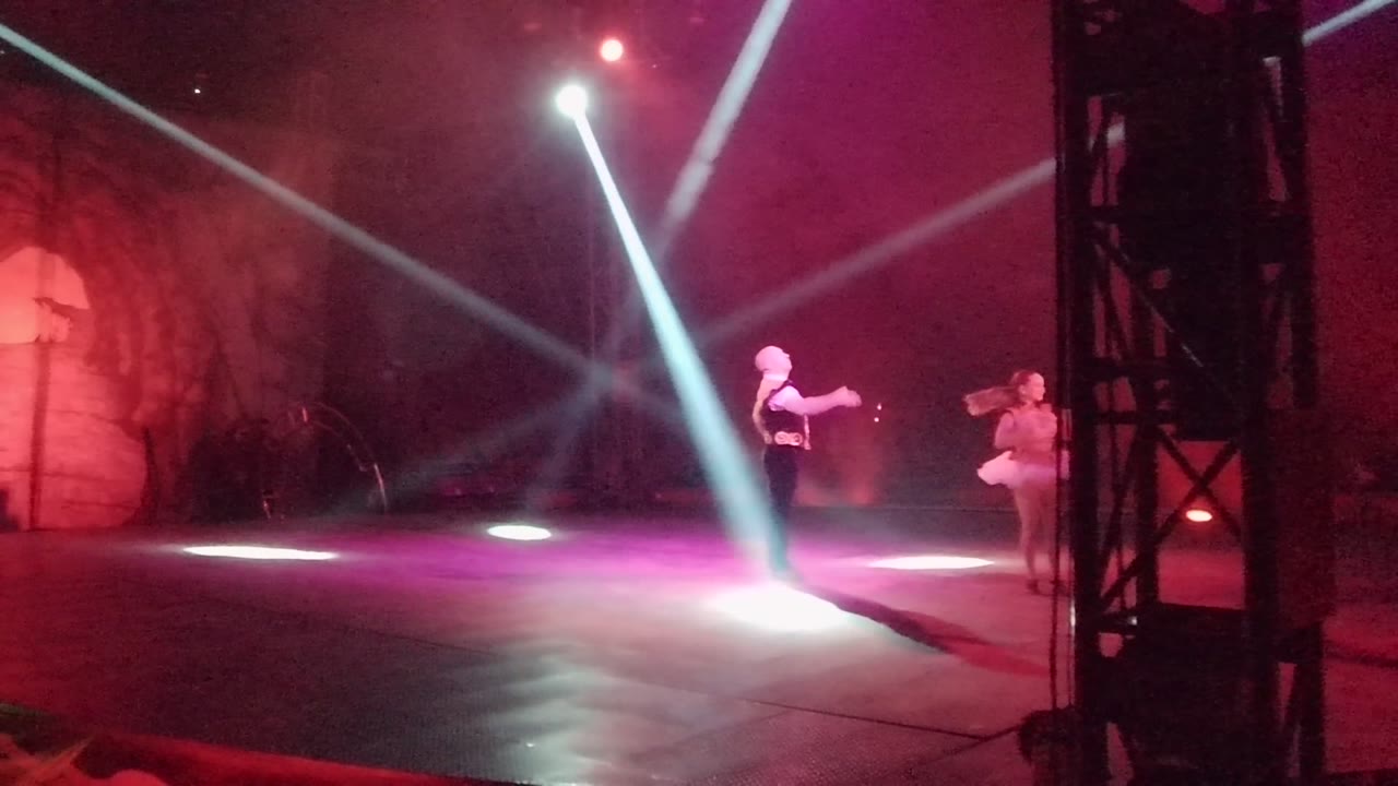 Two stage dancers separated under bright white beams and pink wash.