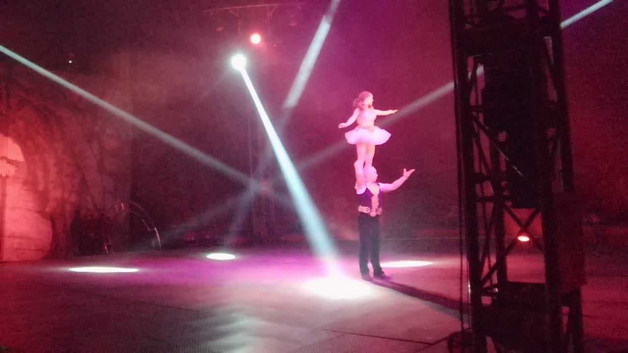In San Jose del Cabo, Mexico, a white-costumed performer balances above a dark-clothed base performer on a pink-lit stage with white spotlights and haze.