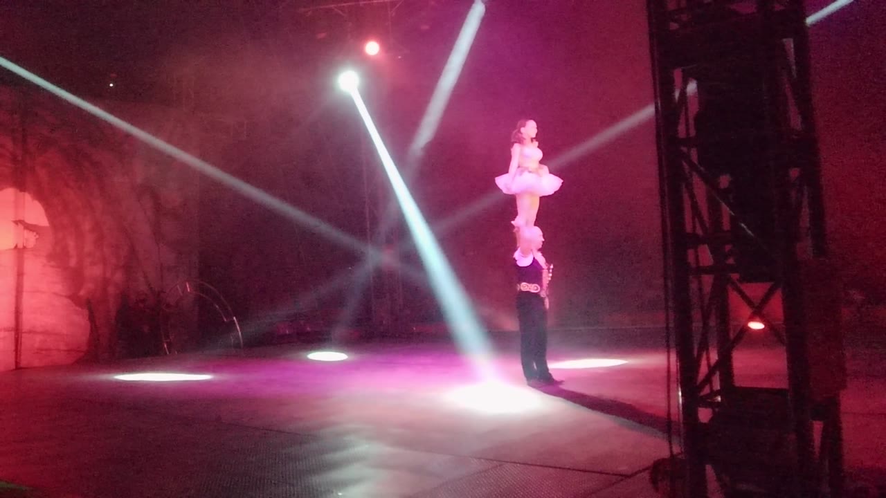 In San Jose del Cabo, Mexico, the white-costumed acrobat reaches a tall elevated pose above the base performer near a bright pool of white stage light.
