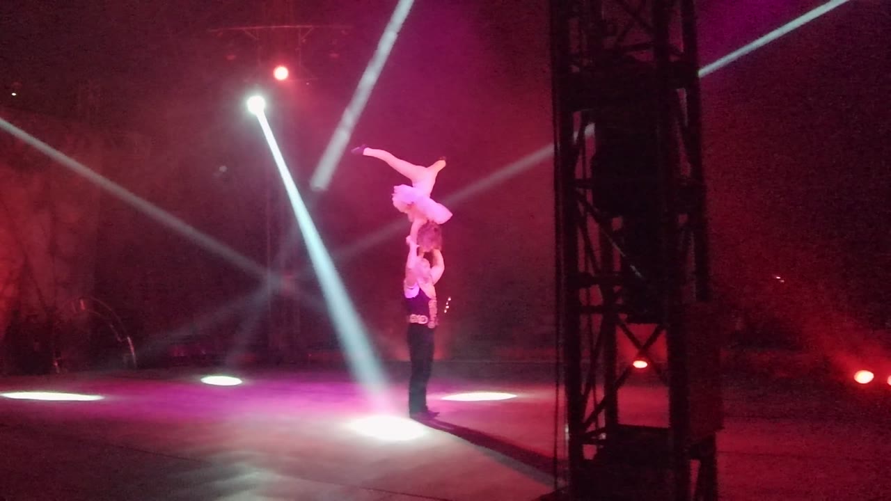 Two acrobats perform a shoulder balance on a dim stage in San Jose Del Cabo BCS Mexico, with the top performer stretched horizontally under pink haze and white spot beams.