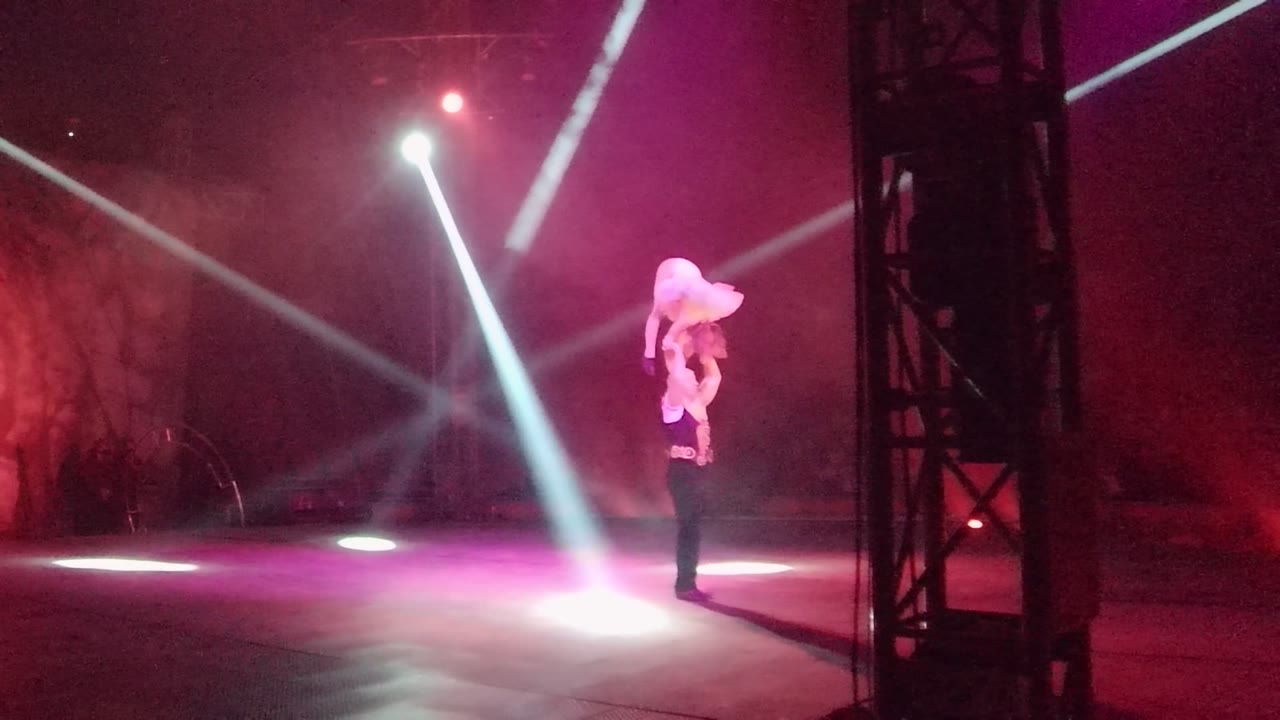 The top performer bends forward while balanced on the base performer in San Jose Del Cabo BCS Mexico, surrounded by pink light, haze, and intersecting white beams.