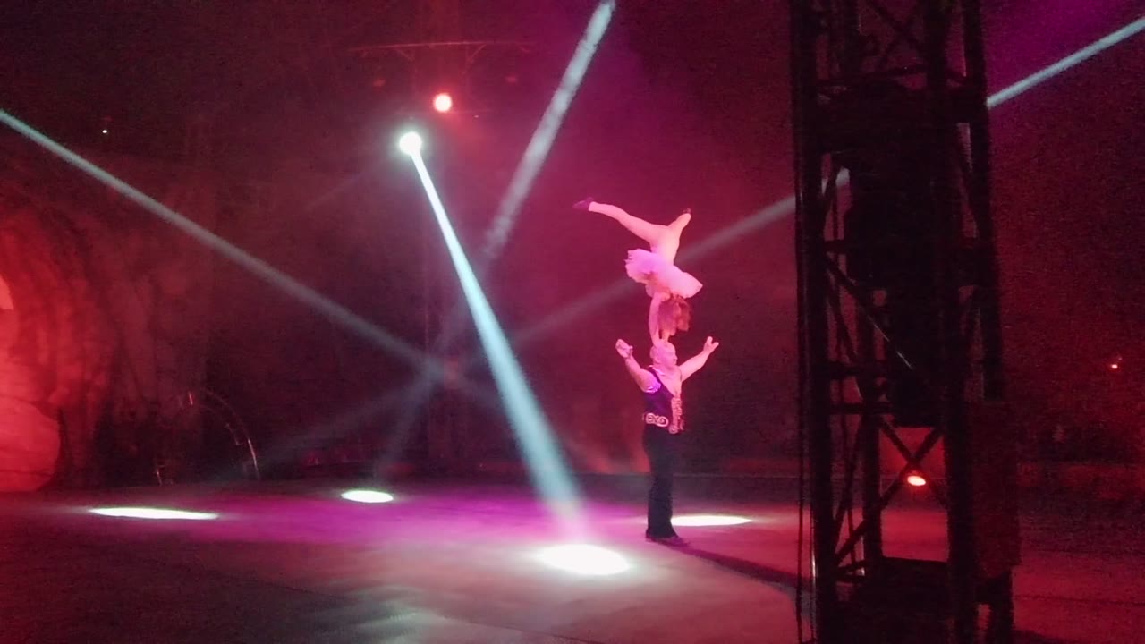 In San Jose del Cabo, Mexico, two acrobats balance on a dim stage as white spotlights cut through red and purple haze.