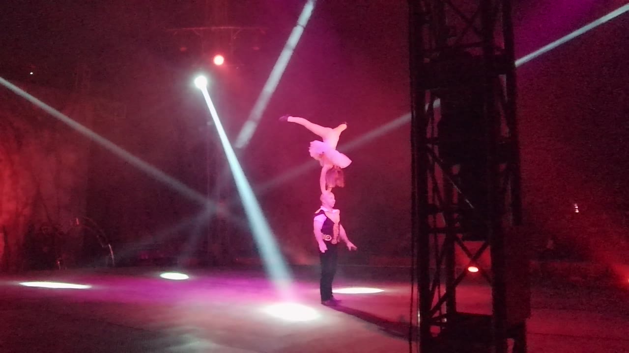 The acrobatic pair holds the balance as the upper performer shifts arm position under red and purple stage lighting in San Jose del Cabo, Mexico.