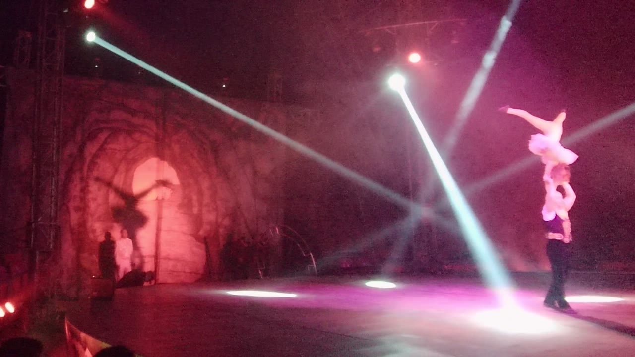 In San Jose Del Cabo BCS Mexico, two acrobats hold a shoulder-lift pose on the right side of a dim stage as white beams cross a red arched backdrop.