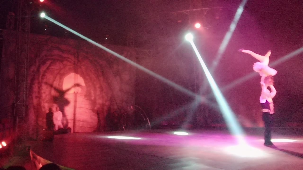 Two acrobats hold a lifted pose while spotlights sweep across a smoky stage.