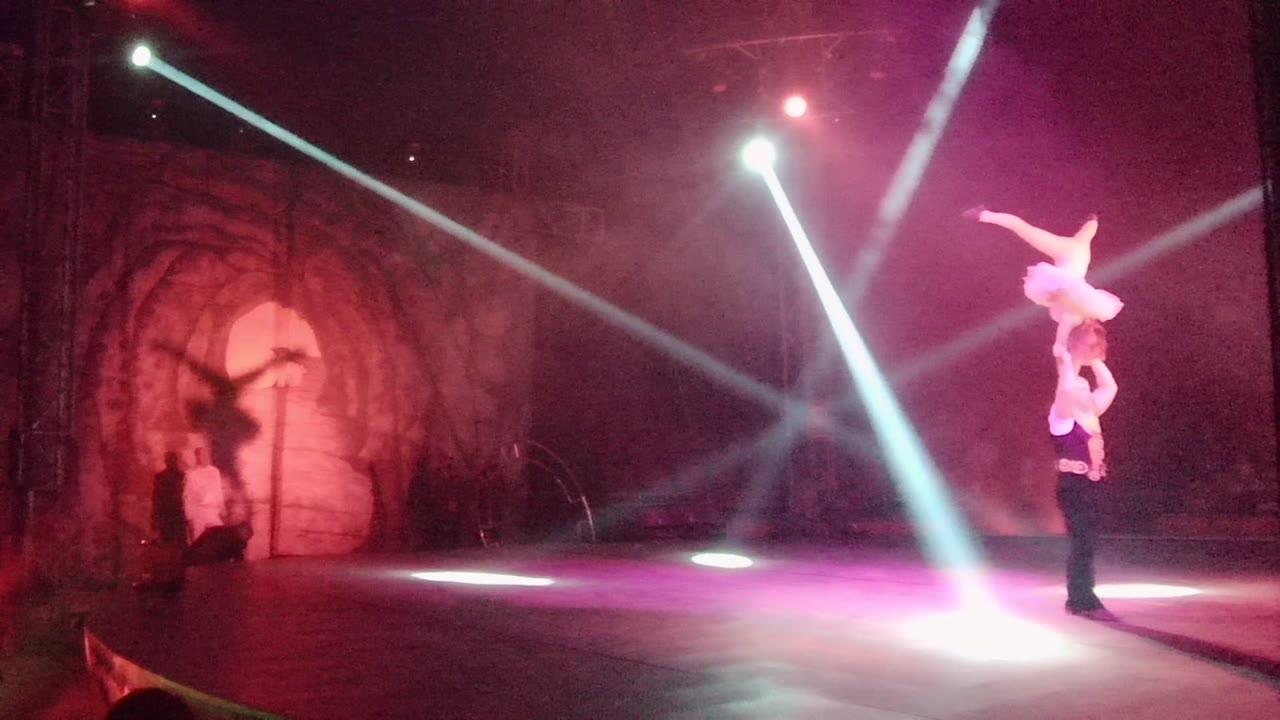 Two performers continue the shoulder balance at the right edge of the stage while strong diagonal beams cross the smoky red and purple scene in San Jose Del Cabo BCS Mexico.