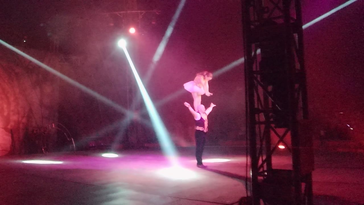 In San Jose del Cabo, Mexico, two performers hold an acrobatic balance on a dark stage while white beams and magenta haze cross behind them.