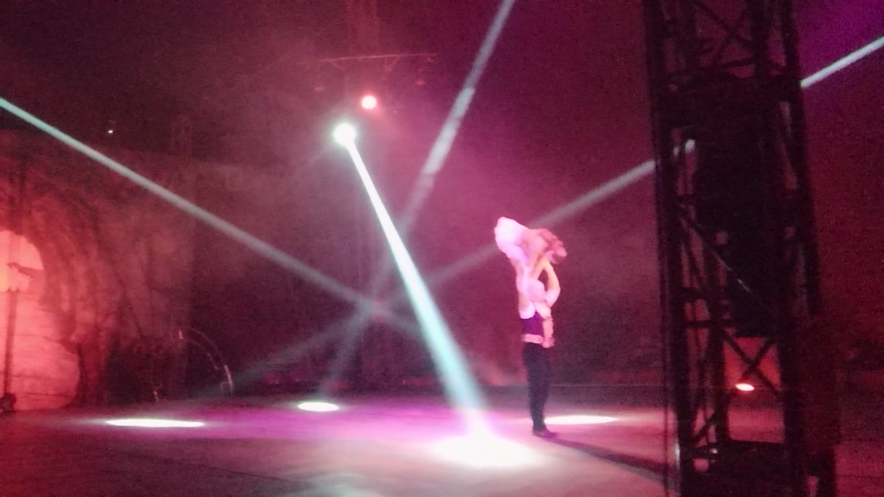 In San Jose del Cabo, Mexico, the lifted performer is held high with legs bent while bright diagonal beams cross over a smoky purple stage.