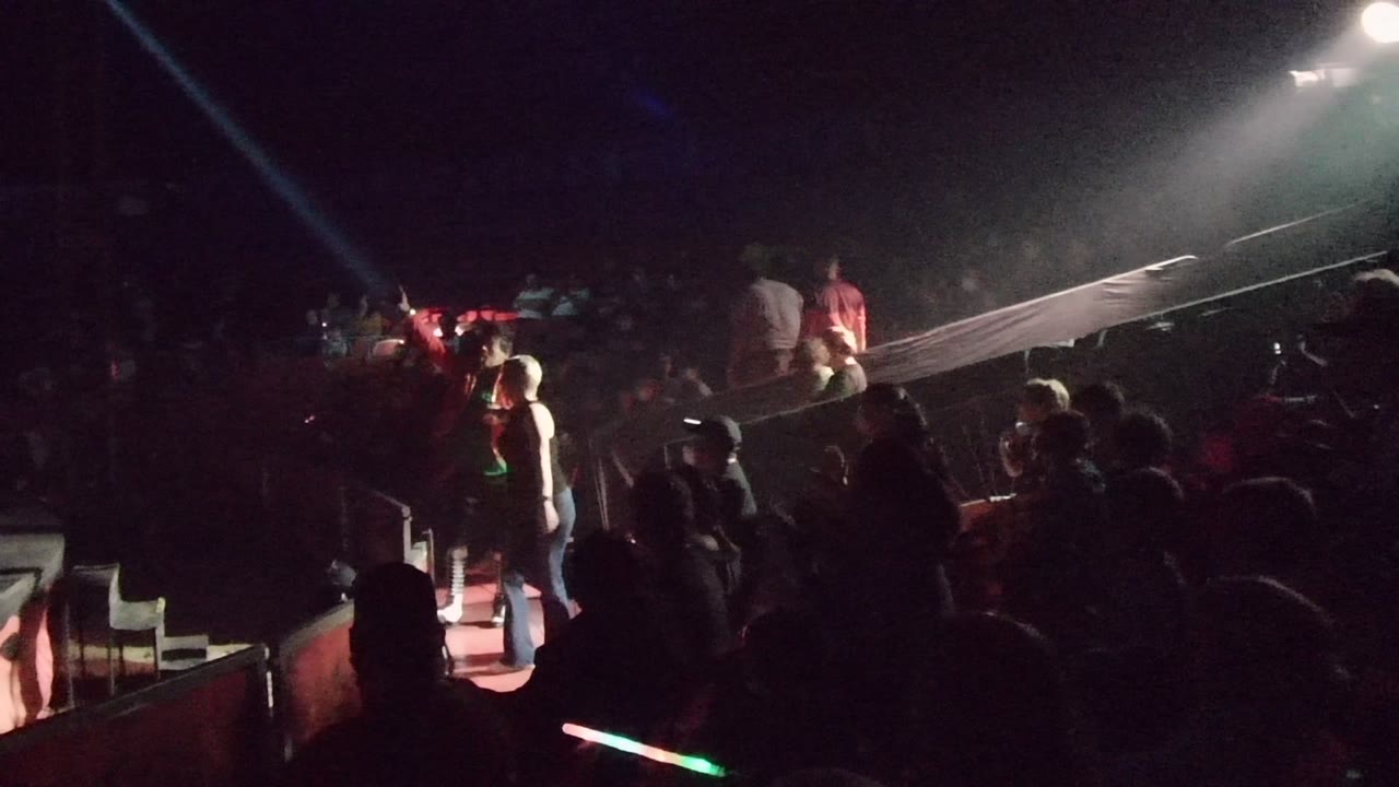 Dim indoor concert scene in San Jose Del Cabo BCS Mexico with performers on a low stage, audience silhouettes in the foreground, red floor light, and a bright overhead spotlight.