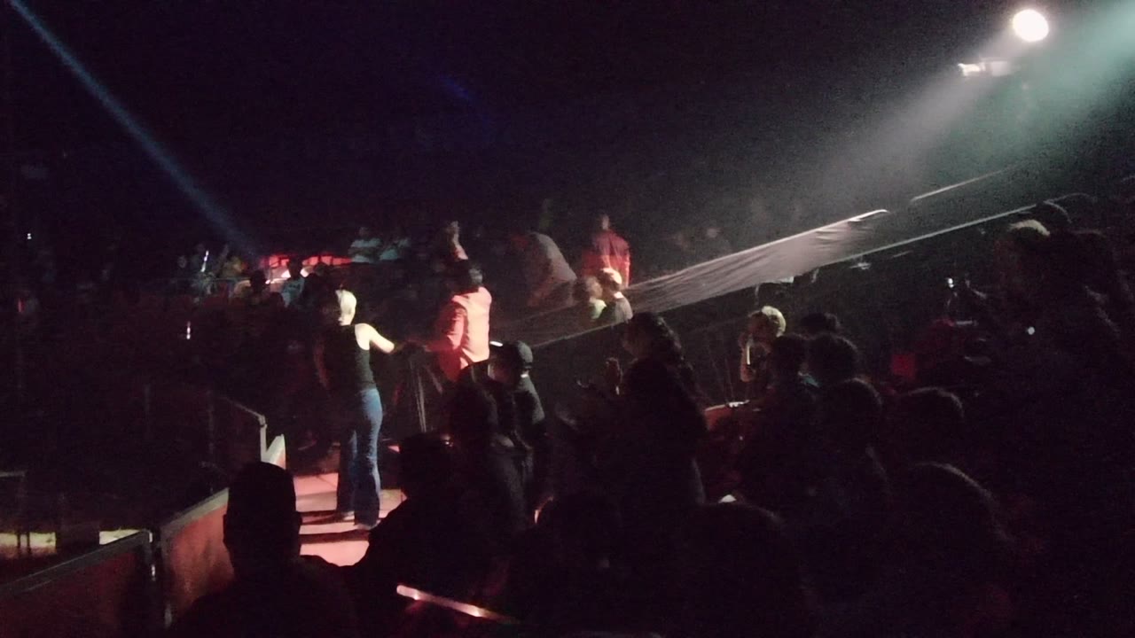 Performers on a dark stage as a spotlight beam crosses the crowd.