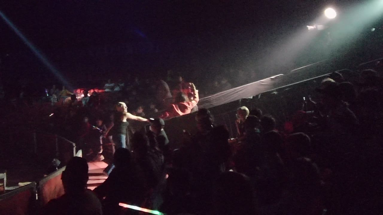 Stage performers remain visible in San Jose Del Cabo BCS Mexico as a bright overhead light pools on the stage and the surrounding audience appears as dark silhouettes with scattered glow sticks.