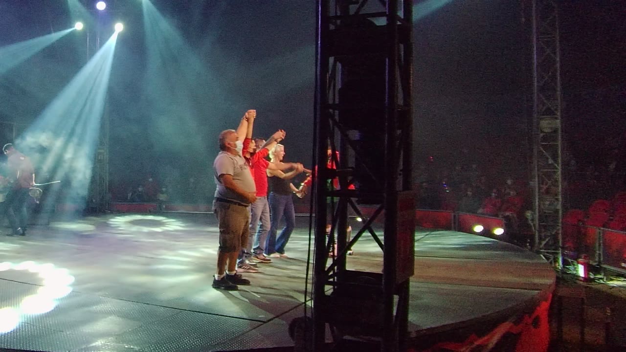 At 0:02 in San Jose del Cabo BCS Mexico, adults stand upright and gesture on the right side of the stage beside a dark truss, with spotlit haze and audience seating in the background.