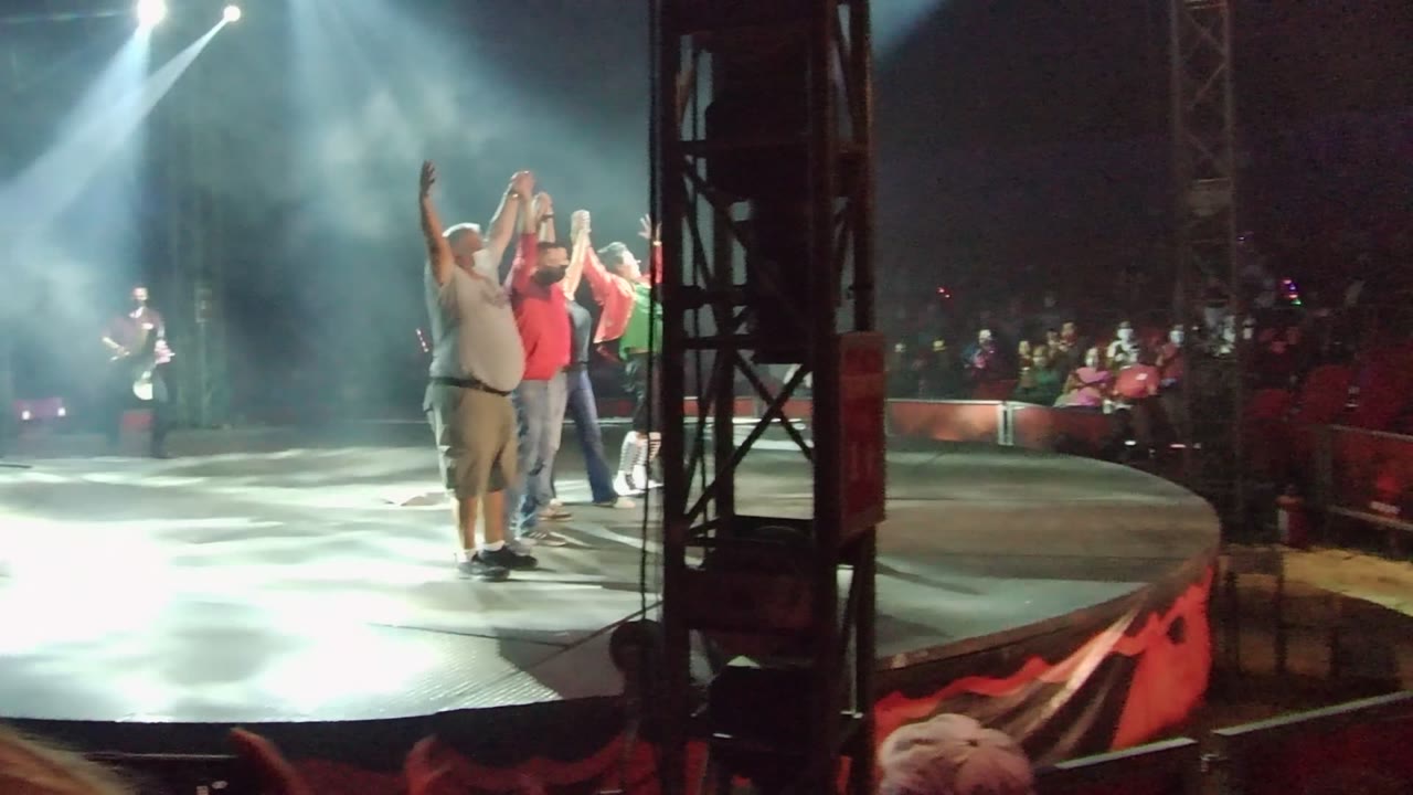 Side-stage view in San Jose Del Cabo BCS Mexico with performers standing near a black truss, some raising arms toward the audience under white beams of light.