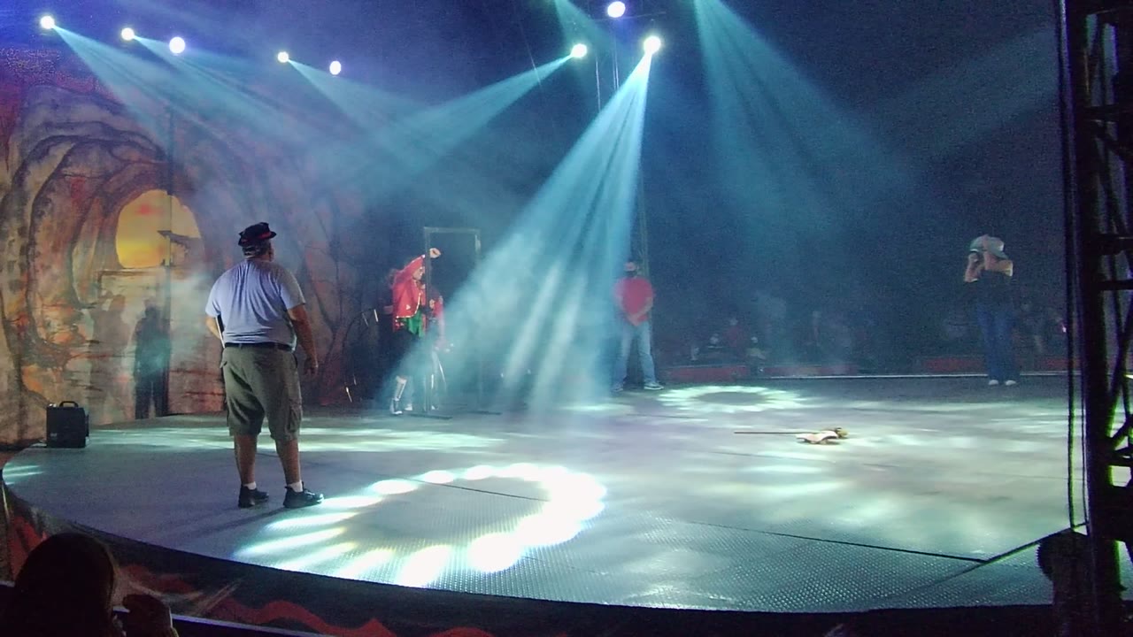 In San Jose del Cabo, Mexico, a wide stage frame shows blue-white spotlights cutting through haze as a red-shirt performer stands midstage and a man in a white shirt and dark hat faces the action from the foreground.