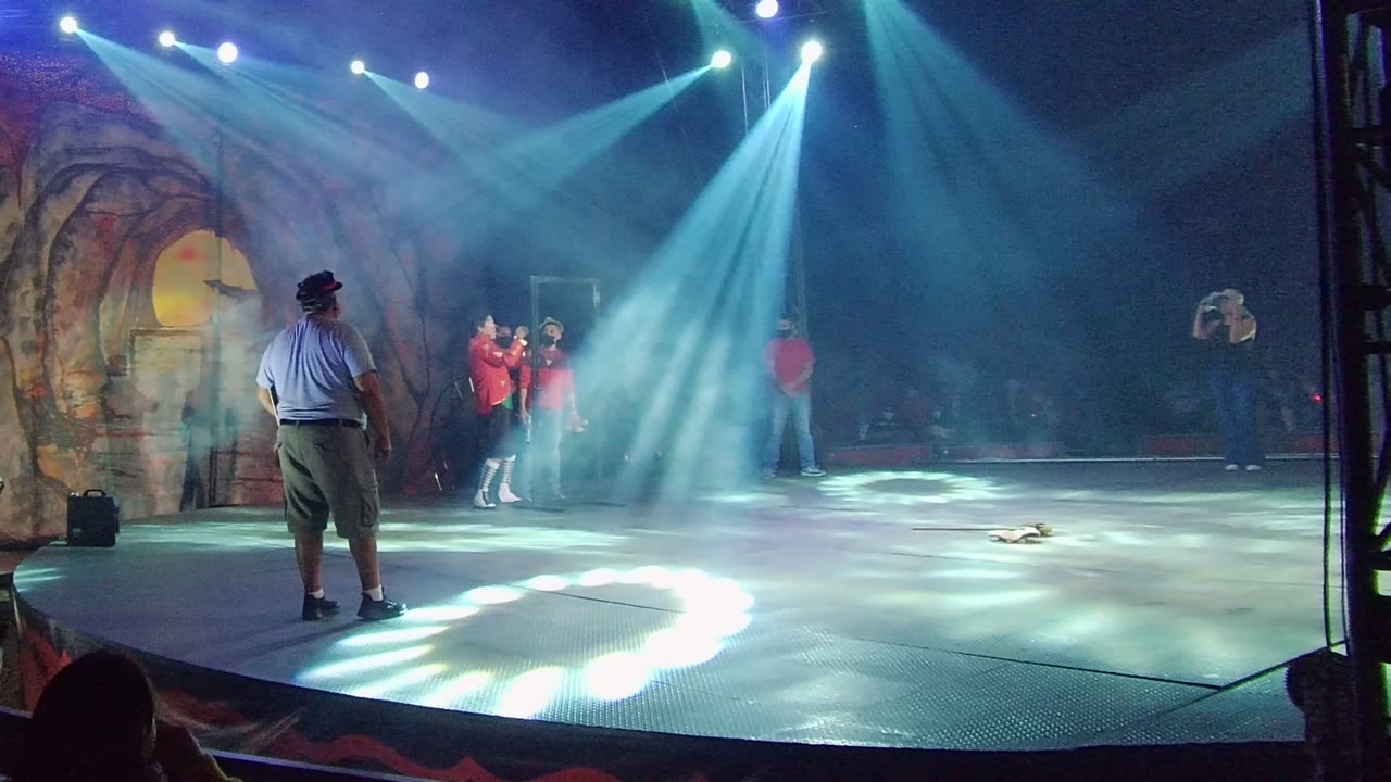 In San Jose del Cabo, Mexico, the red-shirt performer has shifted closer to the center under bright angled spotlights while the foreground man in a hat remains facing the stage.