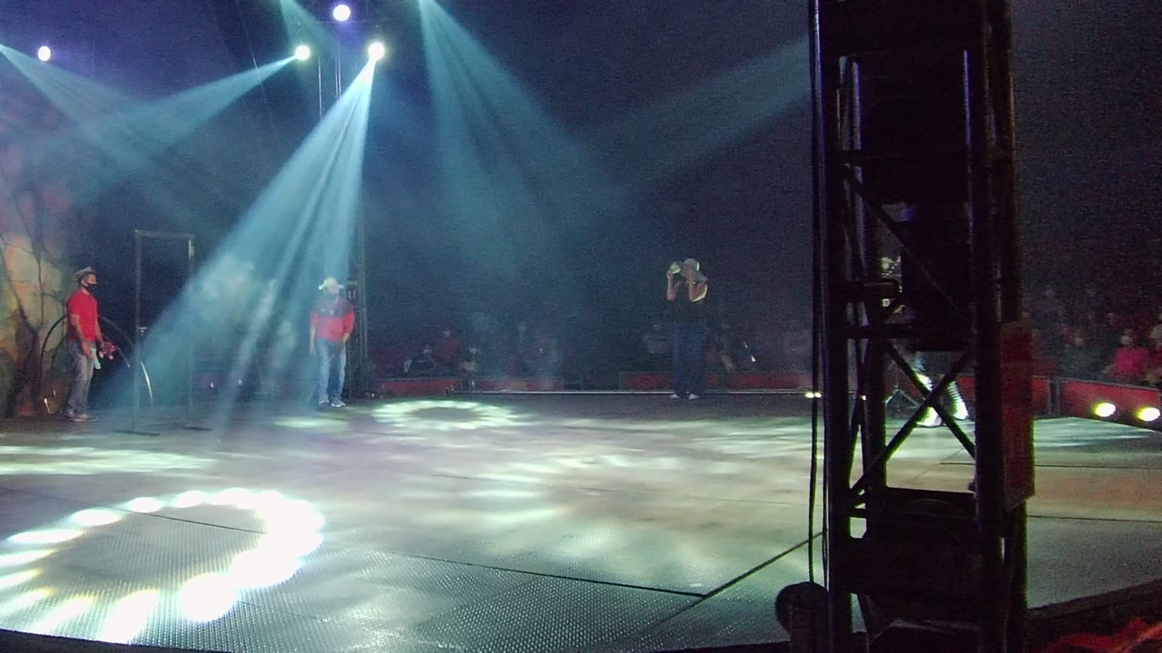 In San Jose del Cabo, Mexico, a wide stage view shows blue-white spotlights crossing the floor while a musician in a red shirt and cowboy hat stands near the left edge with other people visible.