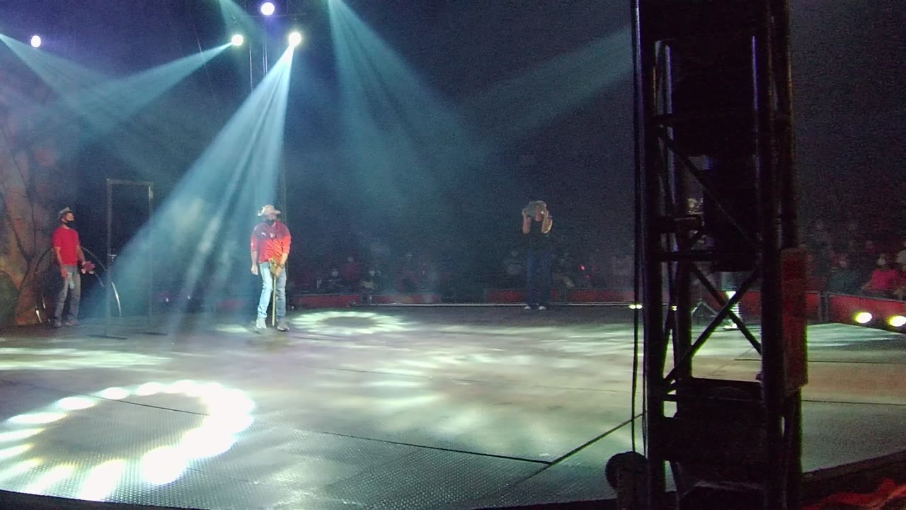 Musician in a red shirt stands nearer center stage under bright overhead beams.