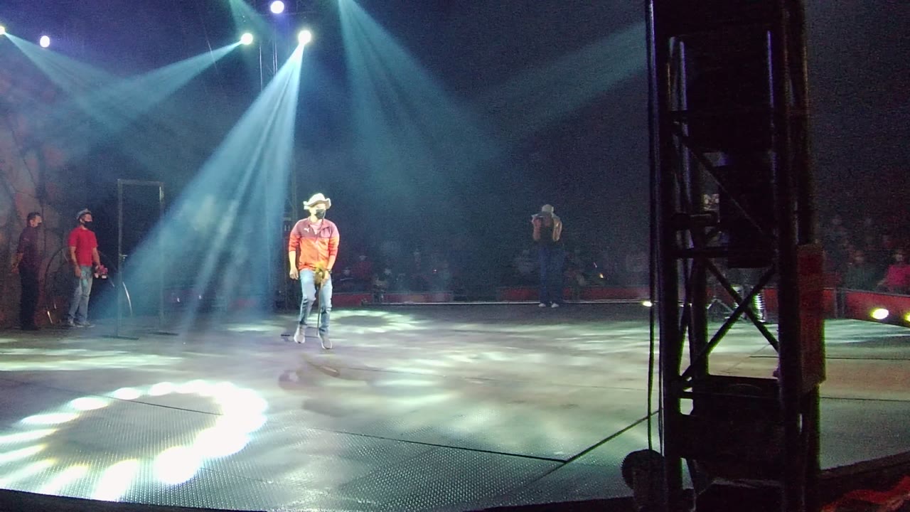 In San Jose del Cabo, Mexico, the guitarist in a red shirt and cowboy hat stands in a bright center-stage pool of light with blue-white beams overhead and audience members visible in the dark background.