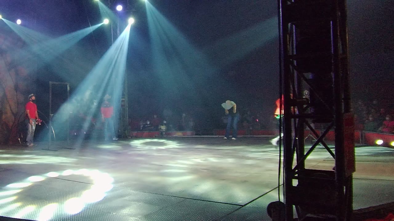 Wide stage performance in San Jose del Cabo, Mexico with blue-white spotlights crossing the floor, a red-shirted performer at left, a white-shirted dancer at right, and audience members behind them.