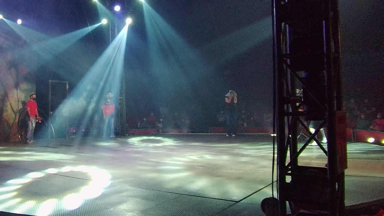 A wide performance frame in San Jose del Cabo, Mexico shows a tall black stage support near the foreground, performers on both sides of the lit stage, and audience seating in the dark background.
