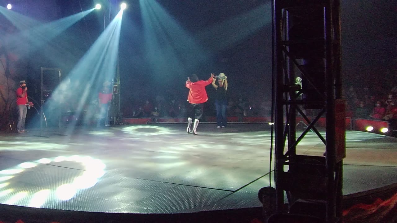 In San Jose del Cabo, Mexico, performers in red shirts stand and move on a stage as blue-white spotlights cross above them and an audience sits in the dark background.