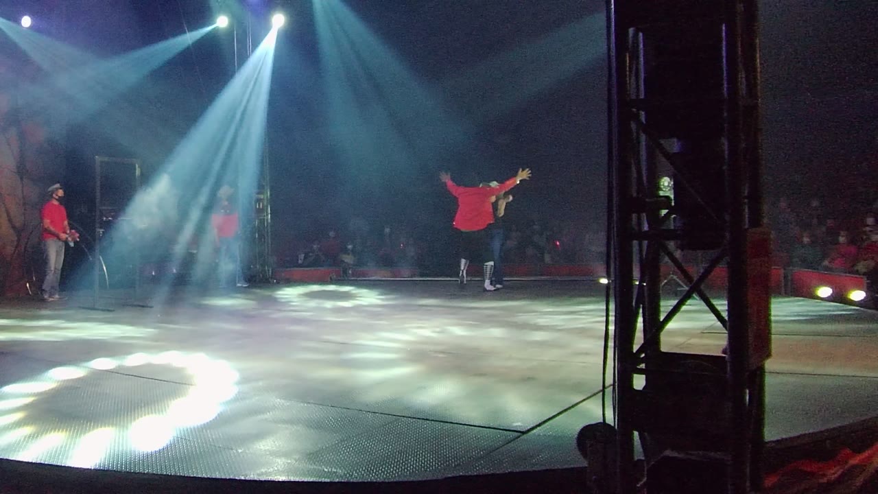 Red-shirt performer gestures near center stage under bright spotlights.