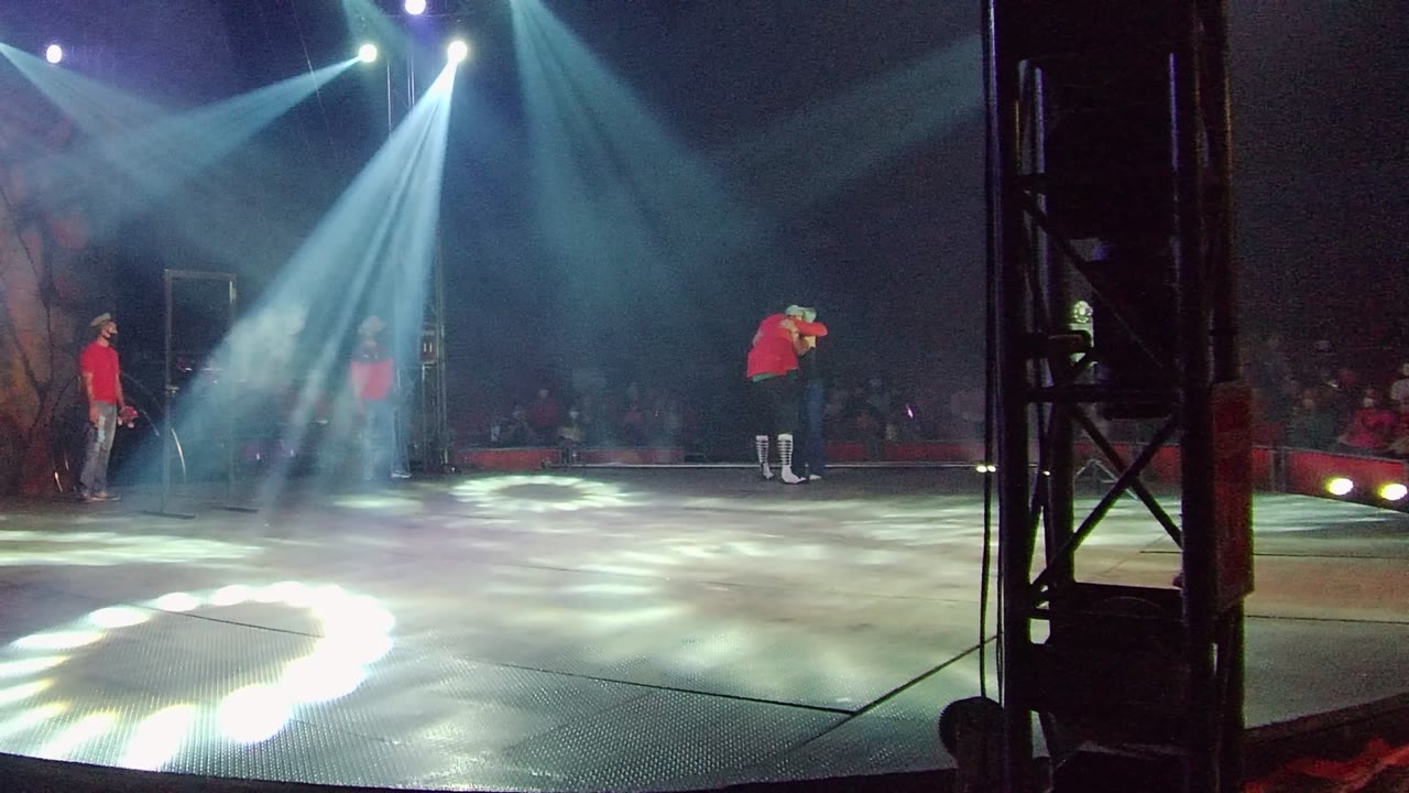 A red-shirt dancer moves across the lit stage in San Jose del Cabo with one arm extended, while blue spotlights and a dark seated audience frame the background.