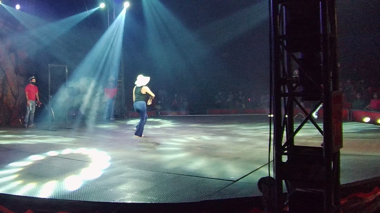 In San Jose del Cabo, Mexico, a dancer in a white cowboy-style hat stands on a blue-white lit stage while another performer in red waits at left and the audience sits in darkness behind them.
