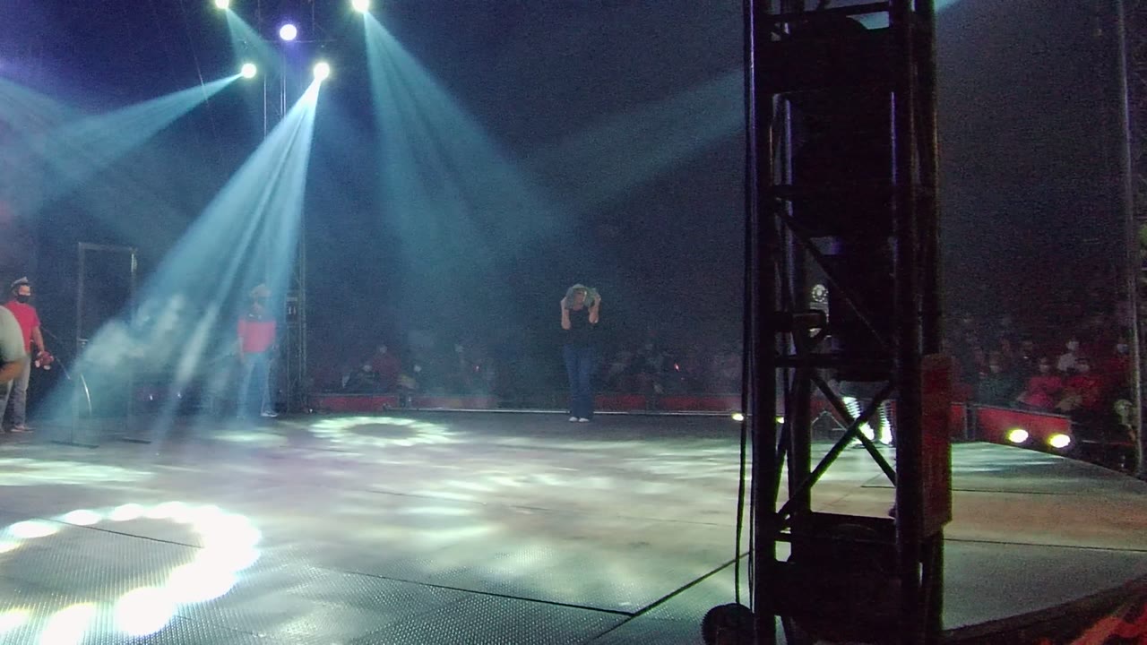 San Jose del Cabo stage performance in Mexico with white-blue spotlights crossing smoke, a red-shirted person near the stage side, another performer at frame edge, and an audience in darkness.