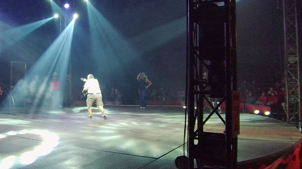 In San Jose del Cabo, Mexico, a dancer in a white shirt stands left of center on a dark stage under blue-white spotlights, with audience members visible in the background.